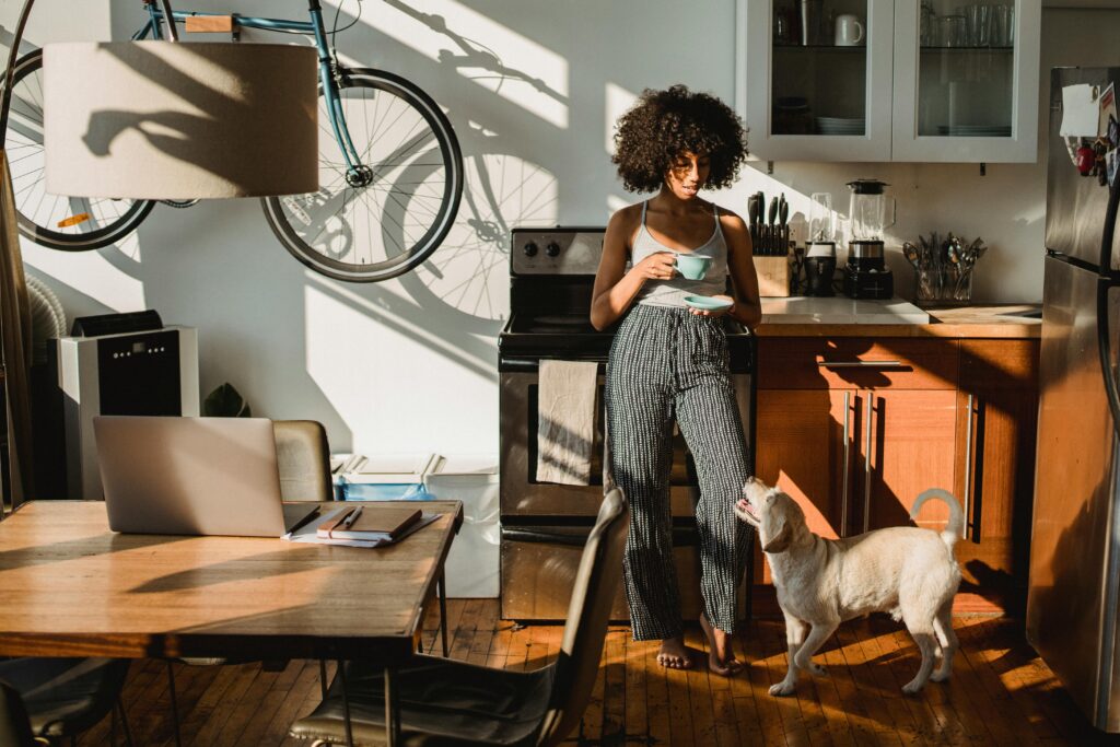 Young barefoot African American female with cup of hot drink standing on floor with dog at home