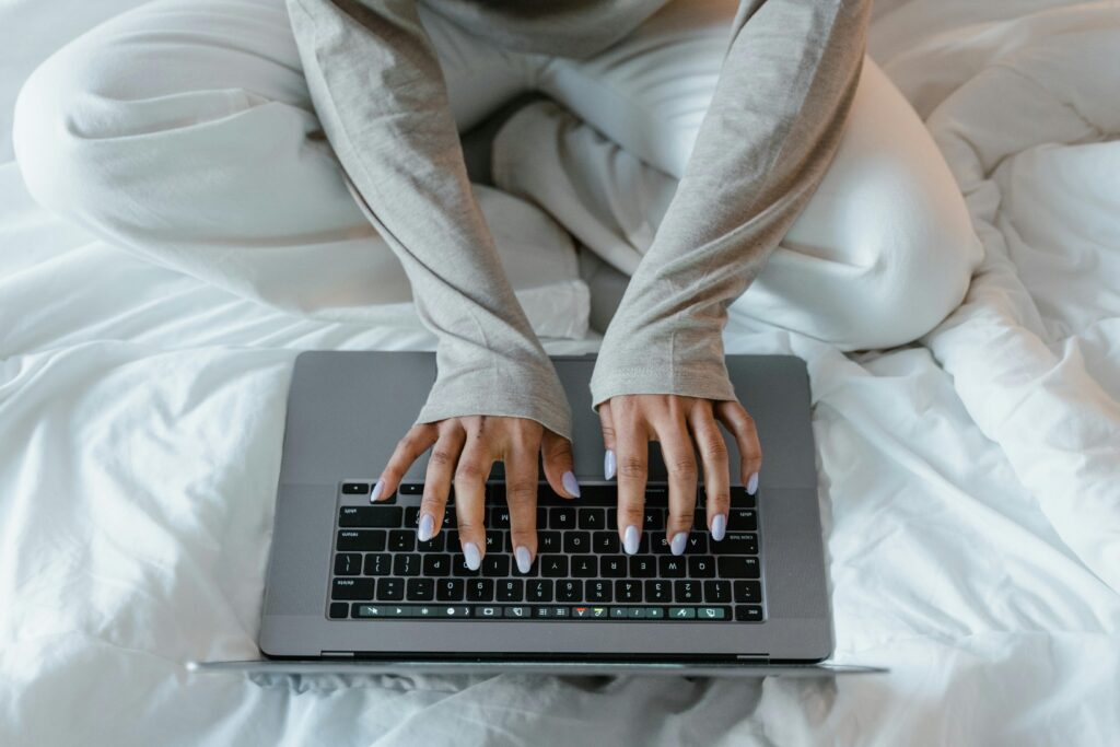 Woman typing on a laptop on a bed, illustrating remote work in a cozy indoor setting.